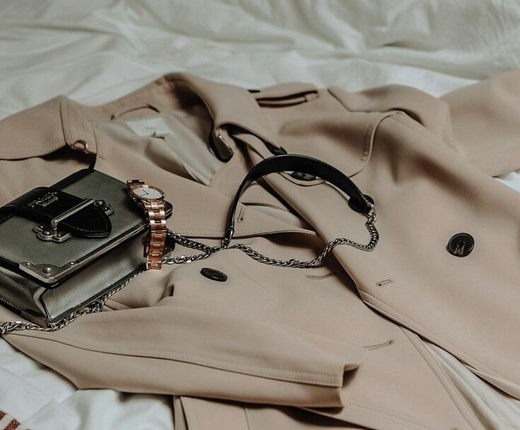 Beige trenchcoat med svart väska och roséguldfärgad klocka på vit säng
