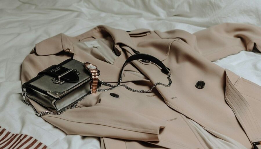 Beige trenchcoat med svart väska och roséguldfärgad klocka på vit säng