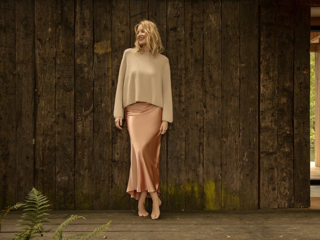 Kvinna i beige stickad tröja och glansig satinkjol mot trävägg